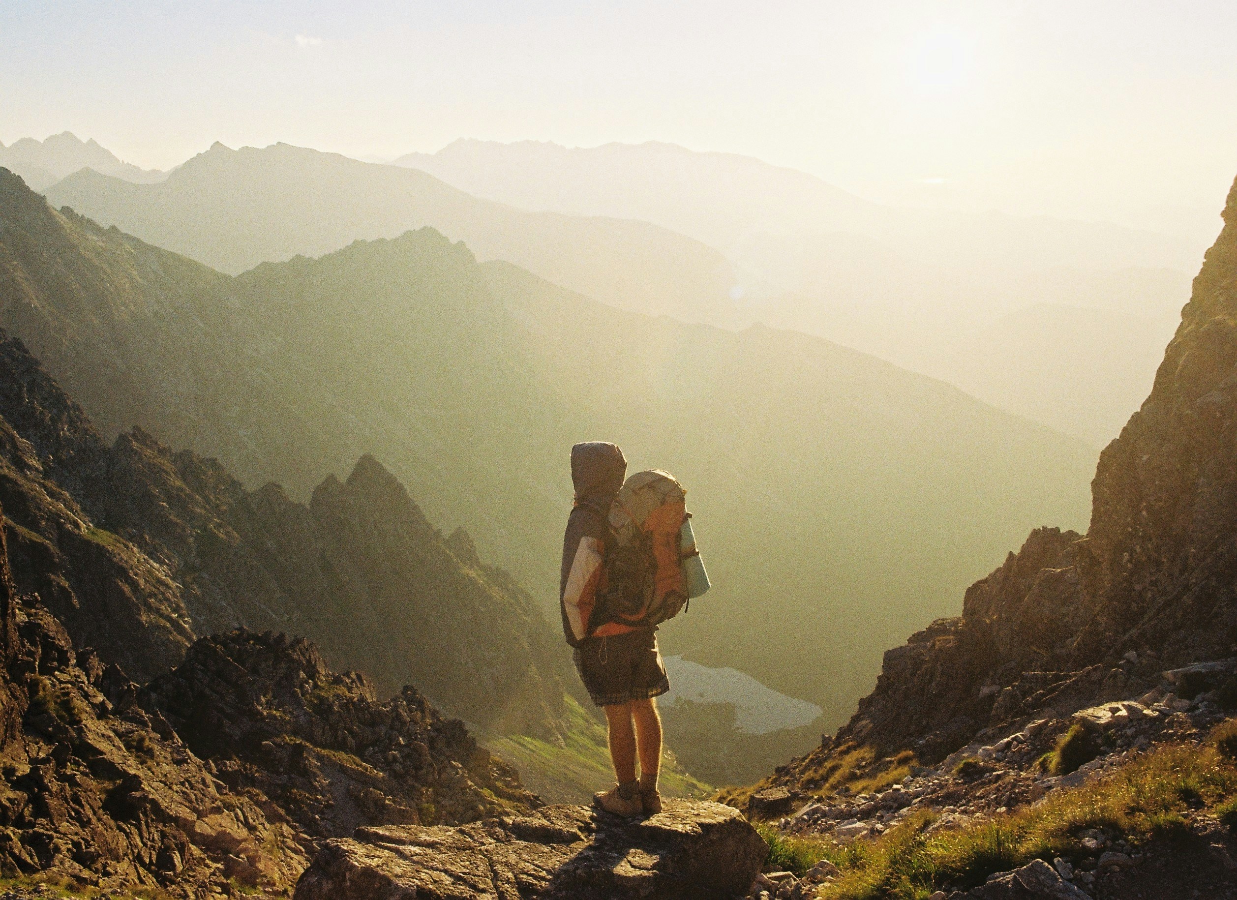 A solo backpacker stands on a rocky mountain ridge at golden hour, looking out over hazy valleys and distant peaks